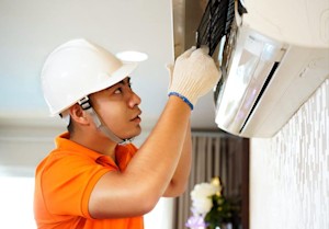 Người thợ mặc áo cam và đội mũ bảo hộ trắng đang sửa máy điều hòa.